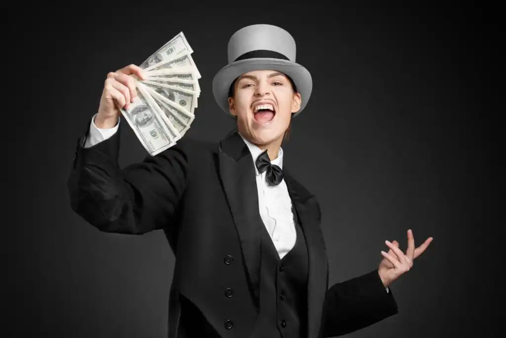 Person in a tuxedo, bow tie, and top hat smiling and holding a fan of U.S. dollar bills against a dark background.