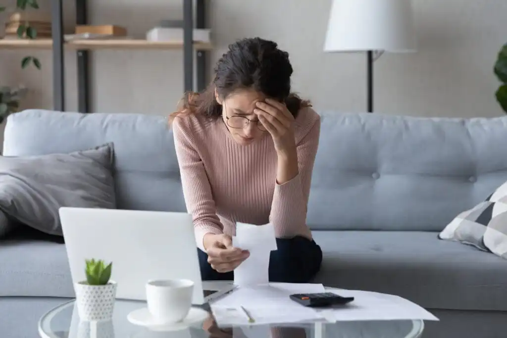 A woman sits on a couch, looking stressed while reviewing paperwork. She holds a piece of paper in one hand and touches her forehead with the other. A laptop, cup, and calculator are on the table in front of her.