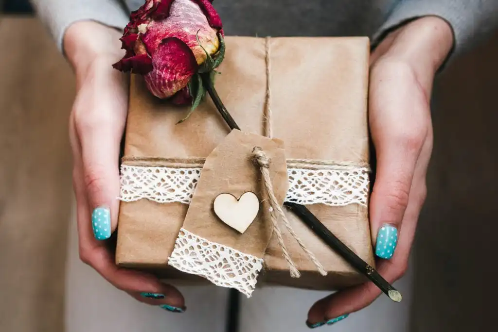 Hands with blue polka dot nails hold a gift wrapped in brown paper, decorated with lace, twine, a dried rose, and a tag with a small wooden heart.