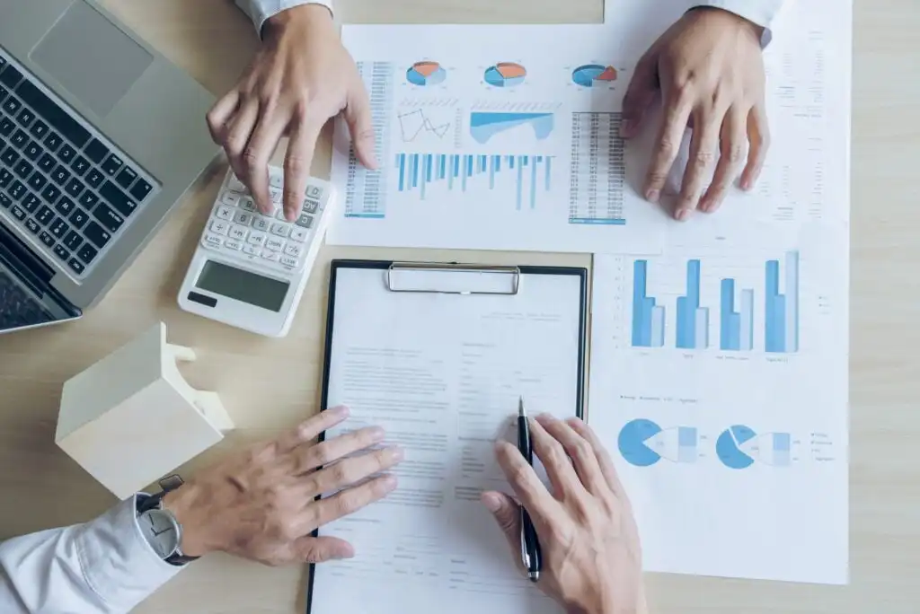 Three people work at a desk with charts, graphs, a clipboard with documents, a calculator, and a laptop, analyzing financial data and discussing business plans.