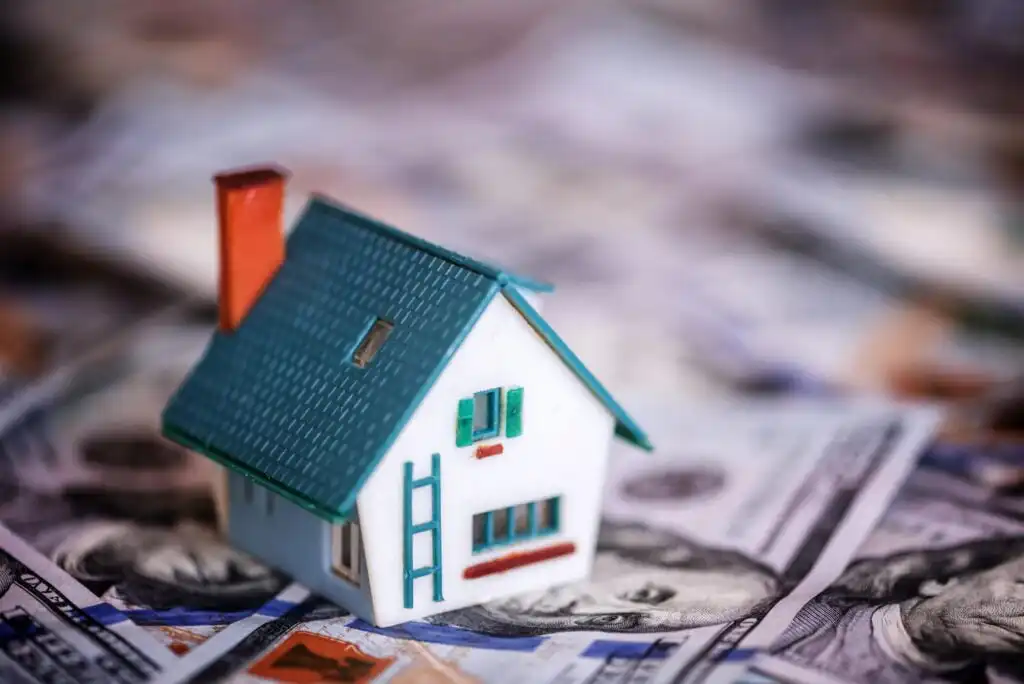 A small model house with a blue roof and green shutters sits on top of several spread-out US hundred-dollar bills, symbolizing real estate investment or housing costs.