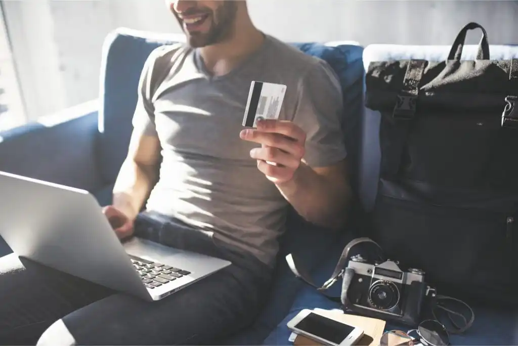 A smiling person sits on a couch using a laptop, holding a credit card. Nearby are a backpack, camera, smartphone, and sunglasses, suggesting travel or online shopping.