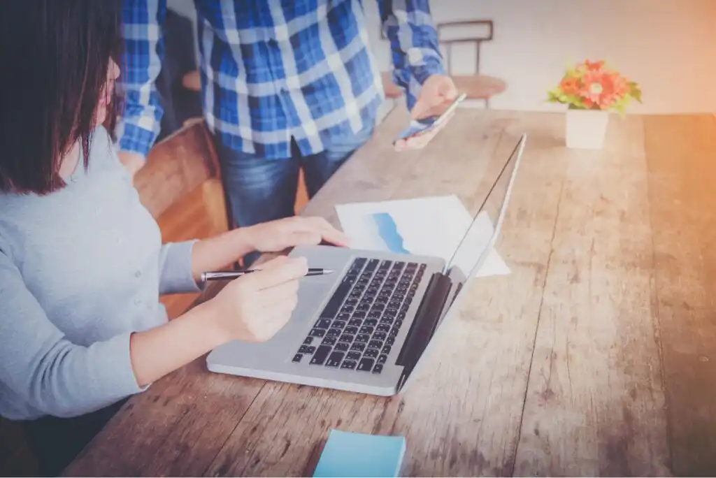Two people working at a wooden table, one typing on a laptop and holding a pen, the other standing nearby holding a smartphone. Papers and a small potted plant are on the table.