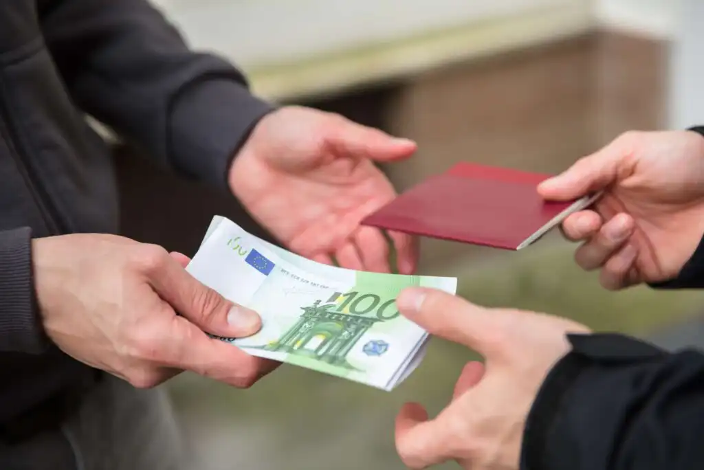Two people exchanging items: one person hands over a stack of 100 euro banknotes, while the other gives a red passport, suggesting a transaction or exchange of money for a passport.