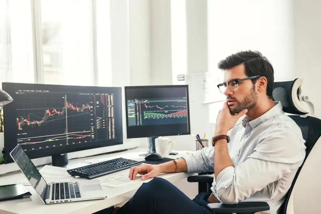 A man wearing glasses sits at a desk with three monitors displaying financial graphs and charts, analyzing data. He looks thoughtful, with one hand on his chin, in a bright, modern office space.
