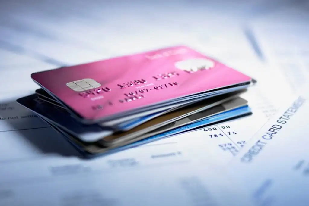 A stack of credit cards, with a pink card on top, lies on several sheets of paper, including a visible credit card statement in the background.