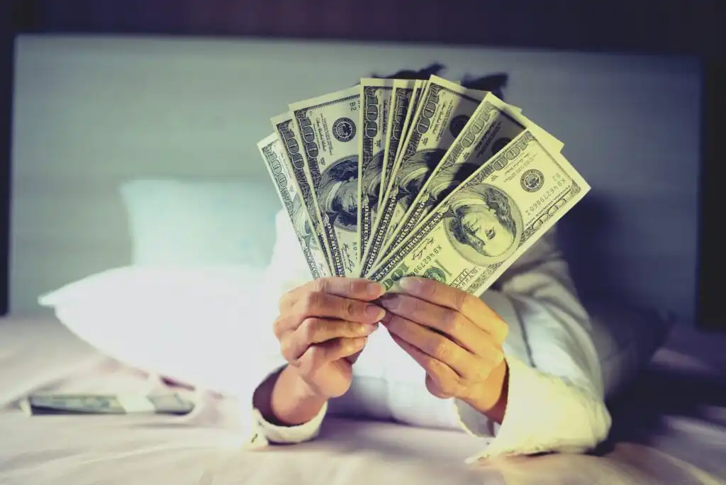 A person sitting on a bed holds and fans out several U.S. hundred-dollar bills, obscuring their face with the money.
