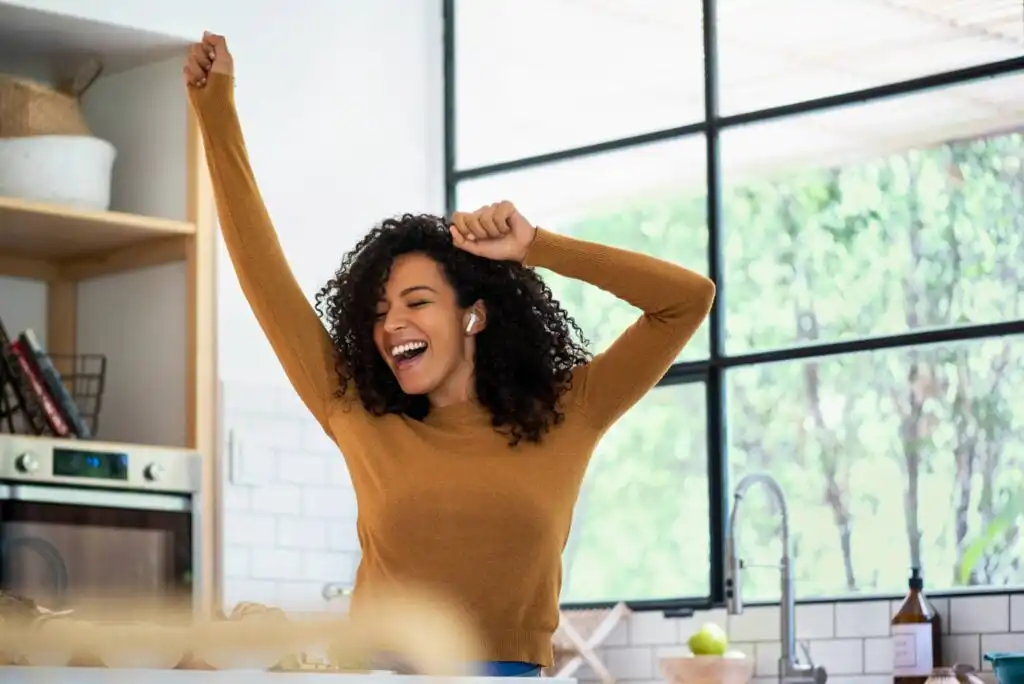 A woman with curly hair wearing a brown sweater and wireless earbuds smiles and raises her arms in excitement while standing in a bright, modern kitchen.