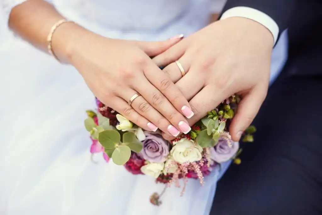 Close-up of a couple’s hands with wedding rings resting on a bouquet of roses and greenery, with the bride in a white dress and the groom in a dark suit.