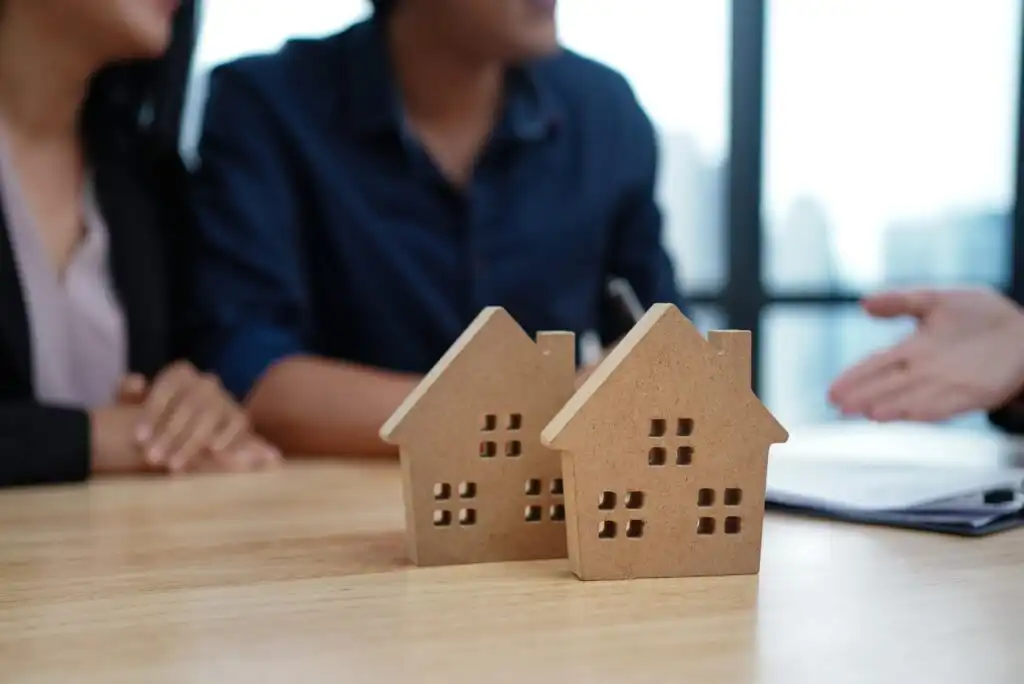 Two wooden house models sit on a table in the foreground. In the background, two people sit together while another person gestures, suggesting a real estate or mortgage discussion.