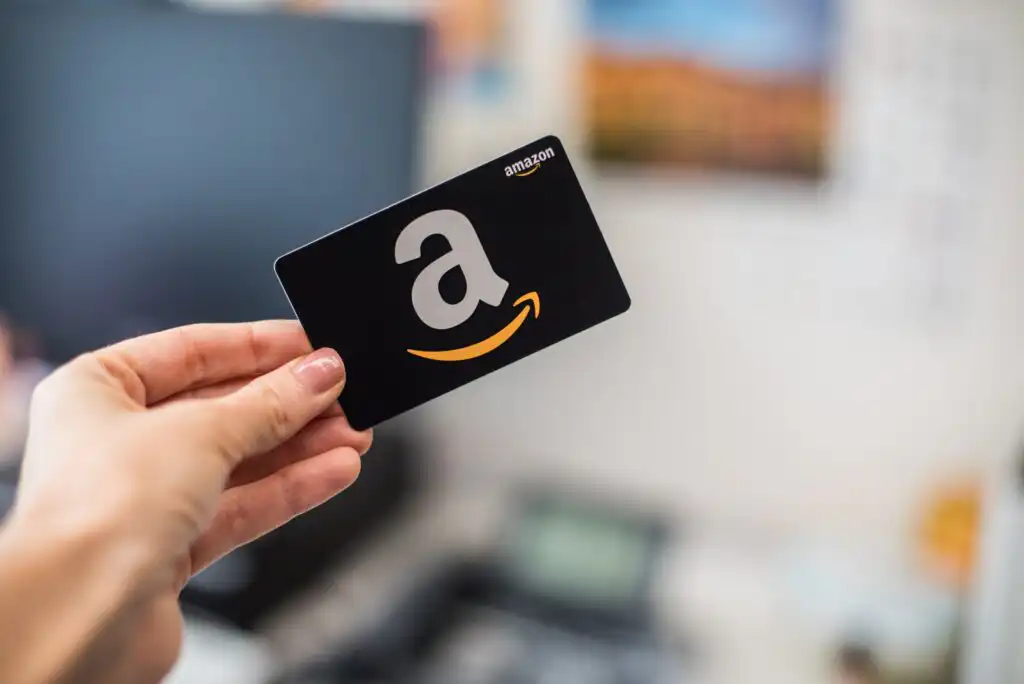 A hand is holding a black Amazon gift card with a large white “a” and a yellow arrow, against a blurred indoor background.