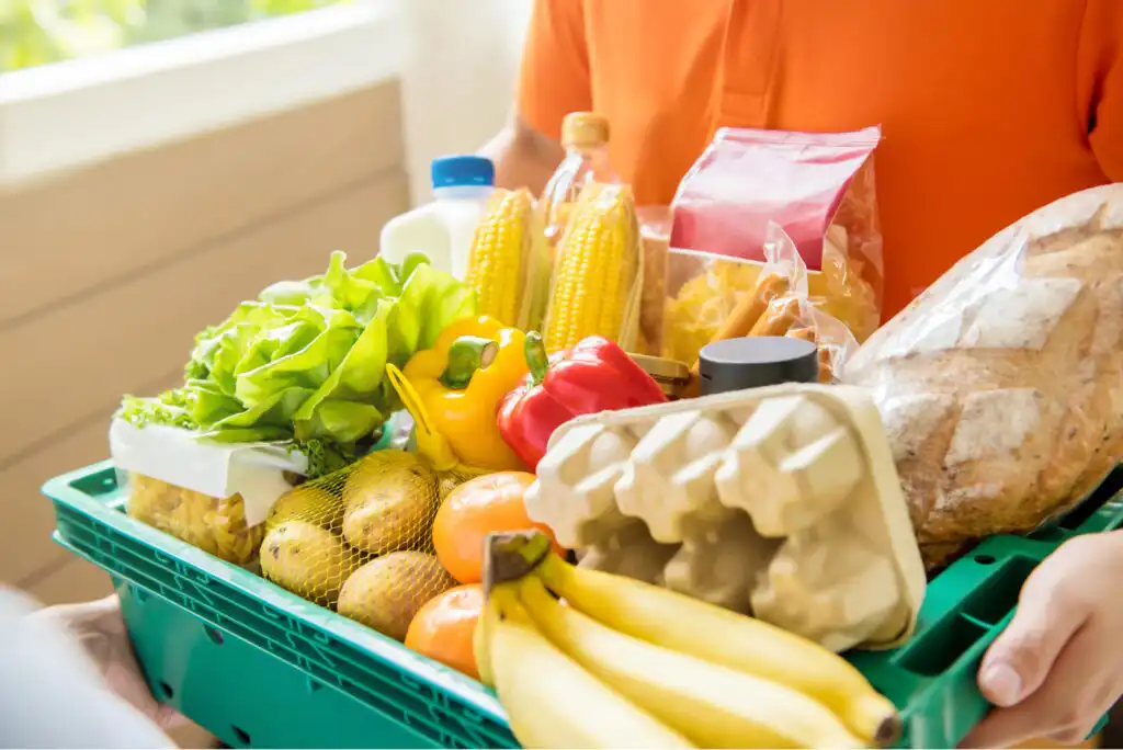A person in an orange shirt holds a green basket filled with groceries, including bananas, bread, eggs, lettuce, bell peppers, potatoes, corn, oranges, pasta, a bottle of oil, and a bottle of milk.