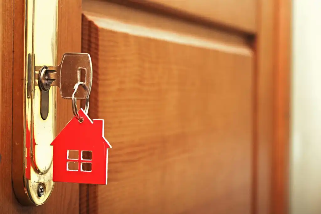 A close-up of a key inserted in a door lock, with a red house-shaped keychain attached, symbolizing home ownership or real estate. The door is wooden with a gold-colored lock plate.