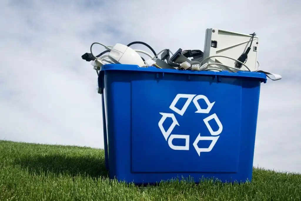 A blue recycling bin filled with various electronic waste, such as cables and old devices, sits on green grass under a partly cloudy sky.
