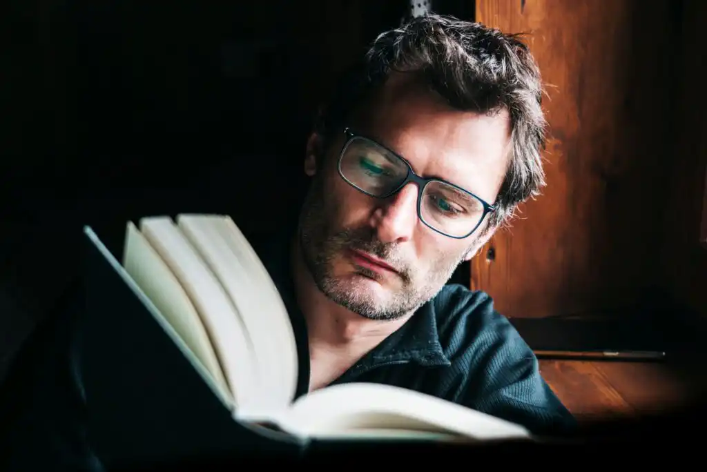 A man with glasses and stubble is reading a large book by a wooden window, illuminated by natural light. He appears focused and thoughtful, with a dark background behind him.
