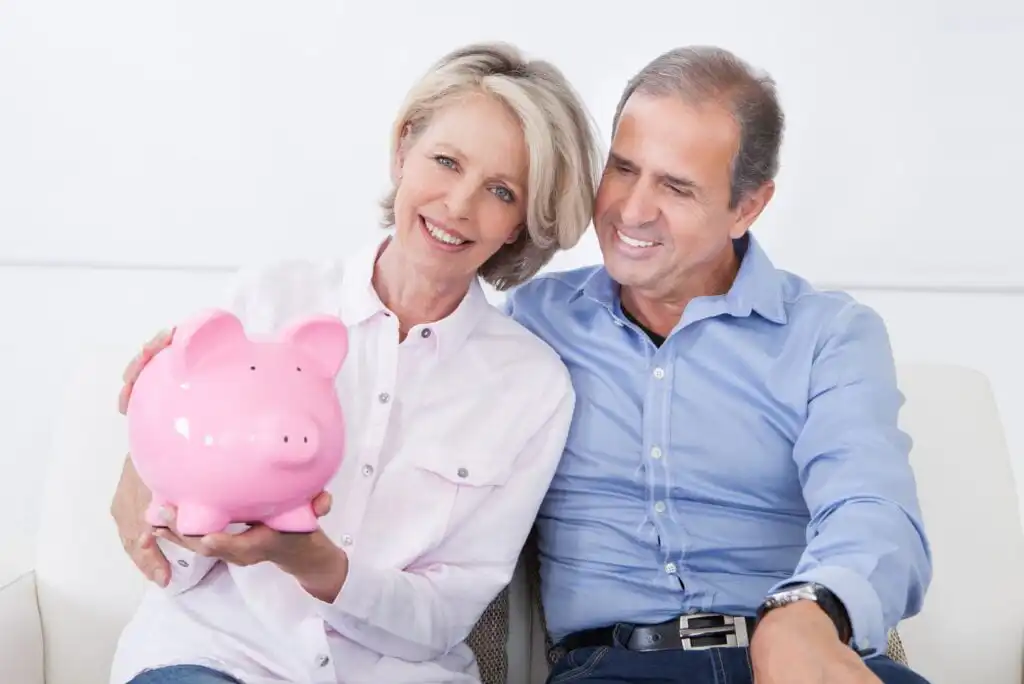 A smiling older couple sits together on a couch. The woman holds a pink piggy bank in her lap, and the man has his arm around her shoulders. Both look happy and relaxed.