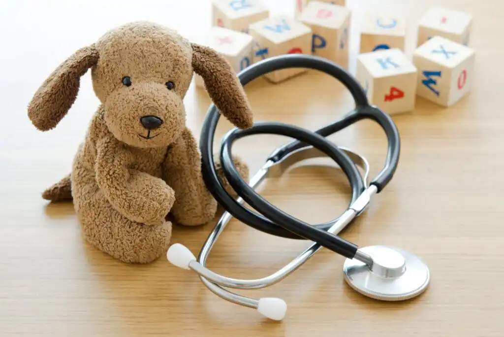 A brown stuffed dog sits next to a stethoscope on a wooden surface, with alphabet blocks in the background.