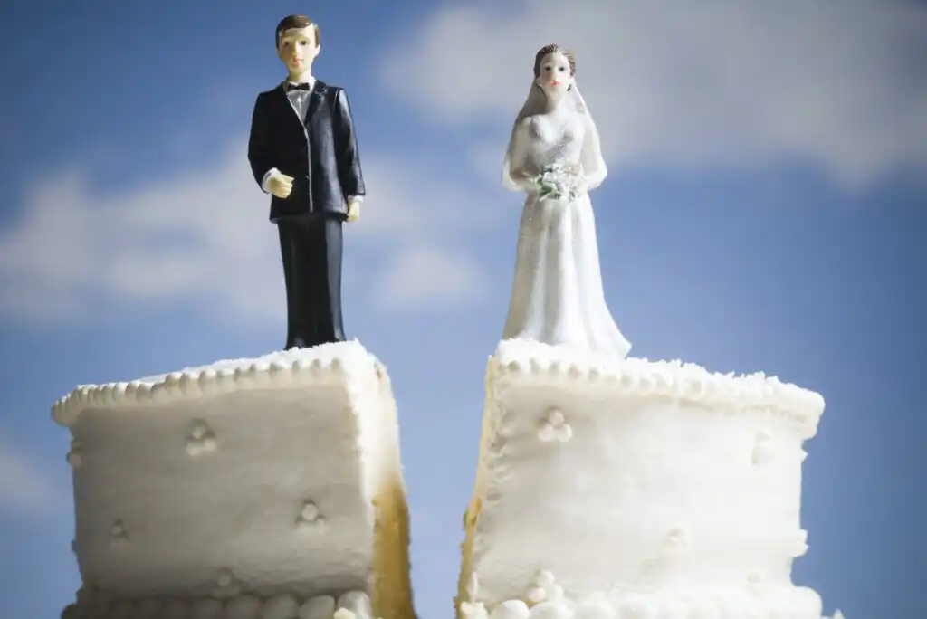 Two wedding cake figurines, a bride and groom, stand on separate pieces of a cake split down the middle, symbolizing separation or divorce, against a blue sky background.