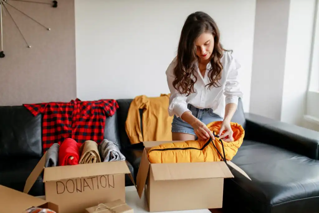 A woman packs clothes into a cardboard box labeled DONATION while standing in a living room, with folded clothes and other boxes on a table in front of her.