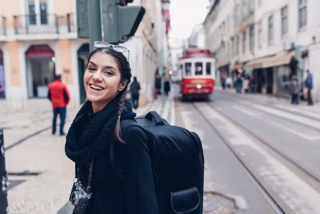 A smiling woman with a backpack and camera stands on a city street near tram tracks, with a red tram approaching in the background. Buildings and pedestrians line the street.