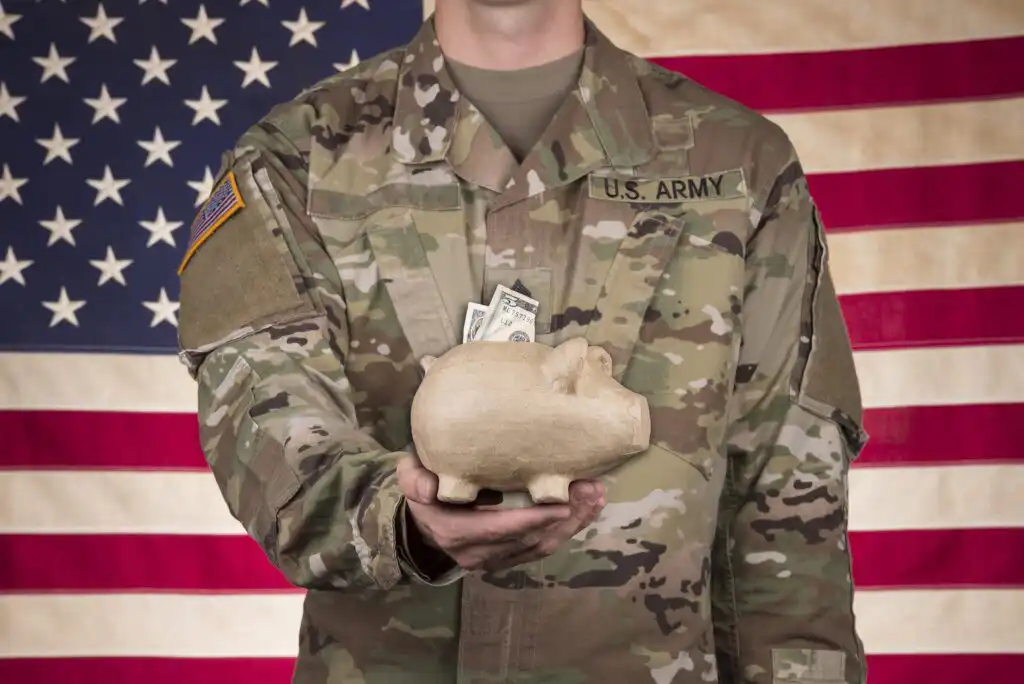 A person in a U.S. Army uniform holds a piggy bank with cash in front of an American flag backdrop.