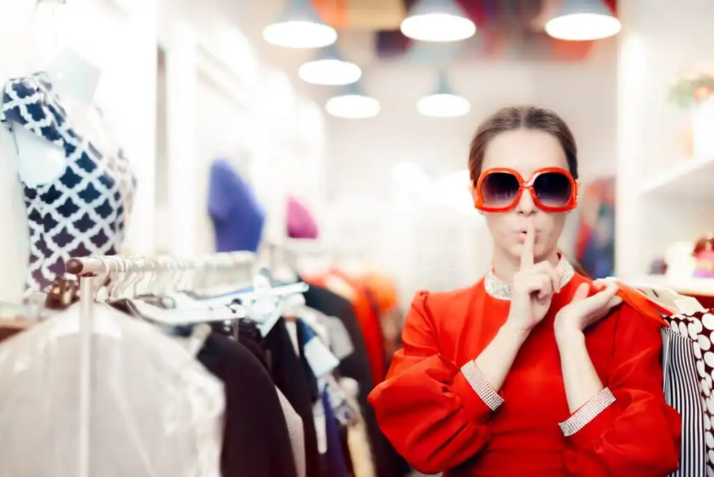 A woman in a bright red dress and oversized red sunglasses stands in a clothing store, holding her finger to her lips in a shushing gesture. Clothing racks and lights are visible in the background.