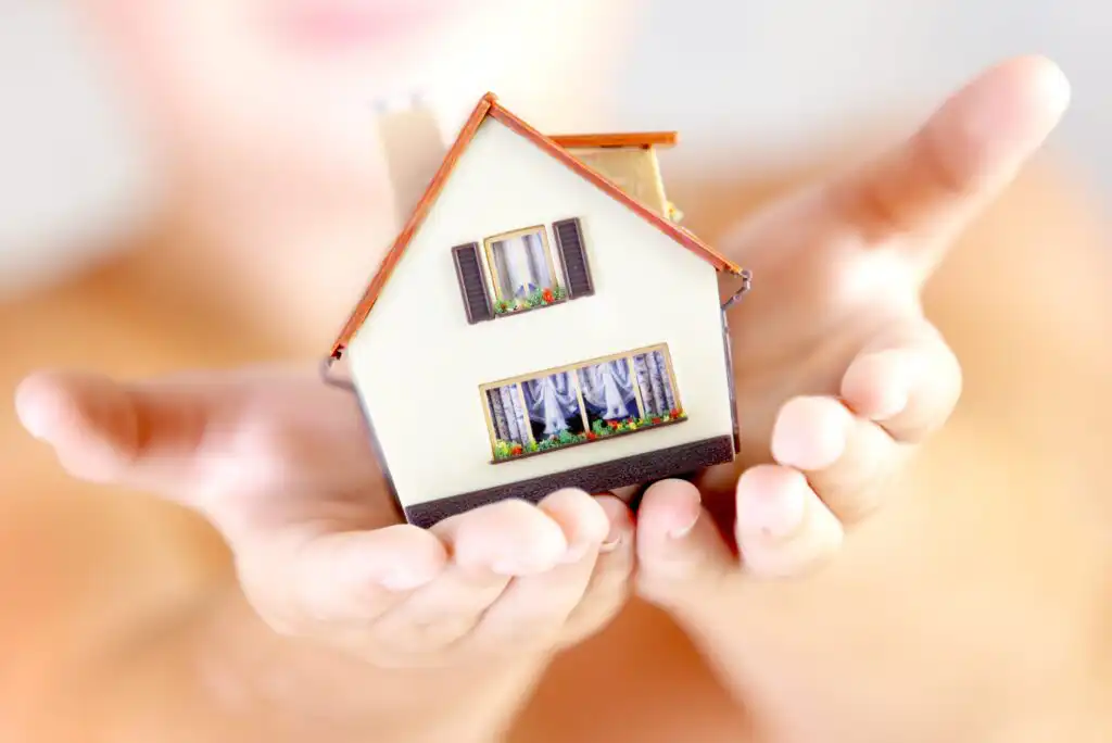 A person holds a small model house with both hands, symbolizing protection, care, or real estate. The background is softly blurred, focusing attention on the detailed miniature house.