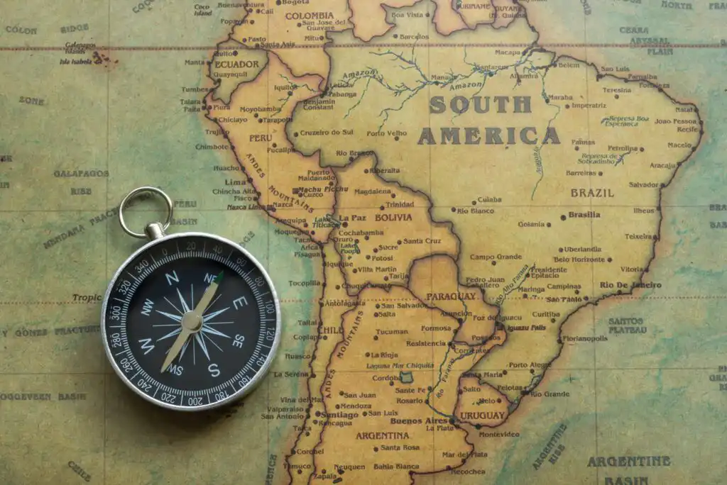 A black and silver compass rests on a vintage map of South America, with country names and borders clearly visible. The compass points northwest, lying over the western part of the continent.